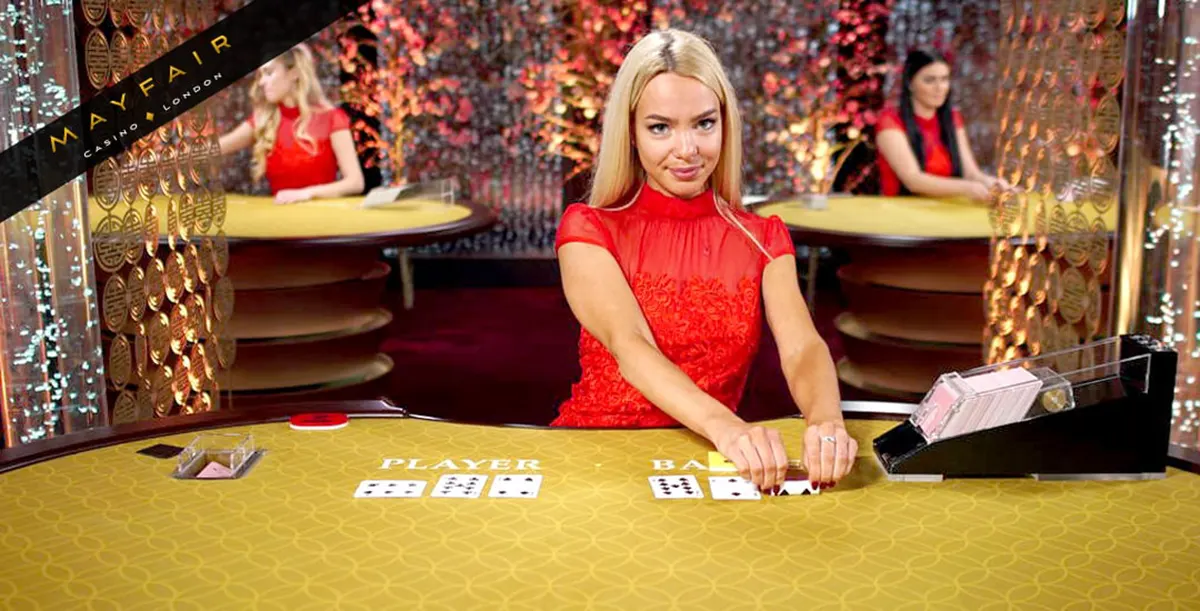Live baccarat scene with a blonde dealer in a red dress dealing Player and Banker hands on a gold felt table, with card shoe and casino-style background featuring other dealers and “Mayfair Casino London” branding.